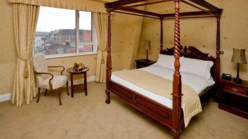 Una cama con dosel de madera, una mesita redonda con una lámpara, una silla y vistas a edificios a través de una ventana con cortinas.