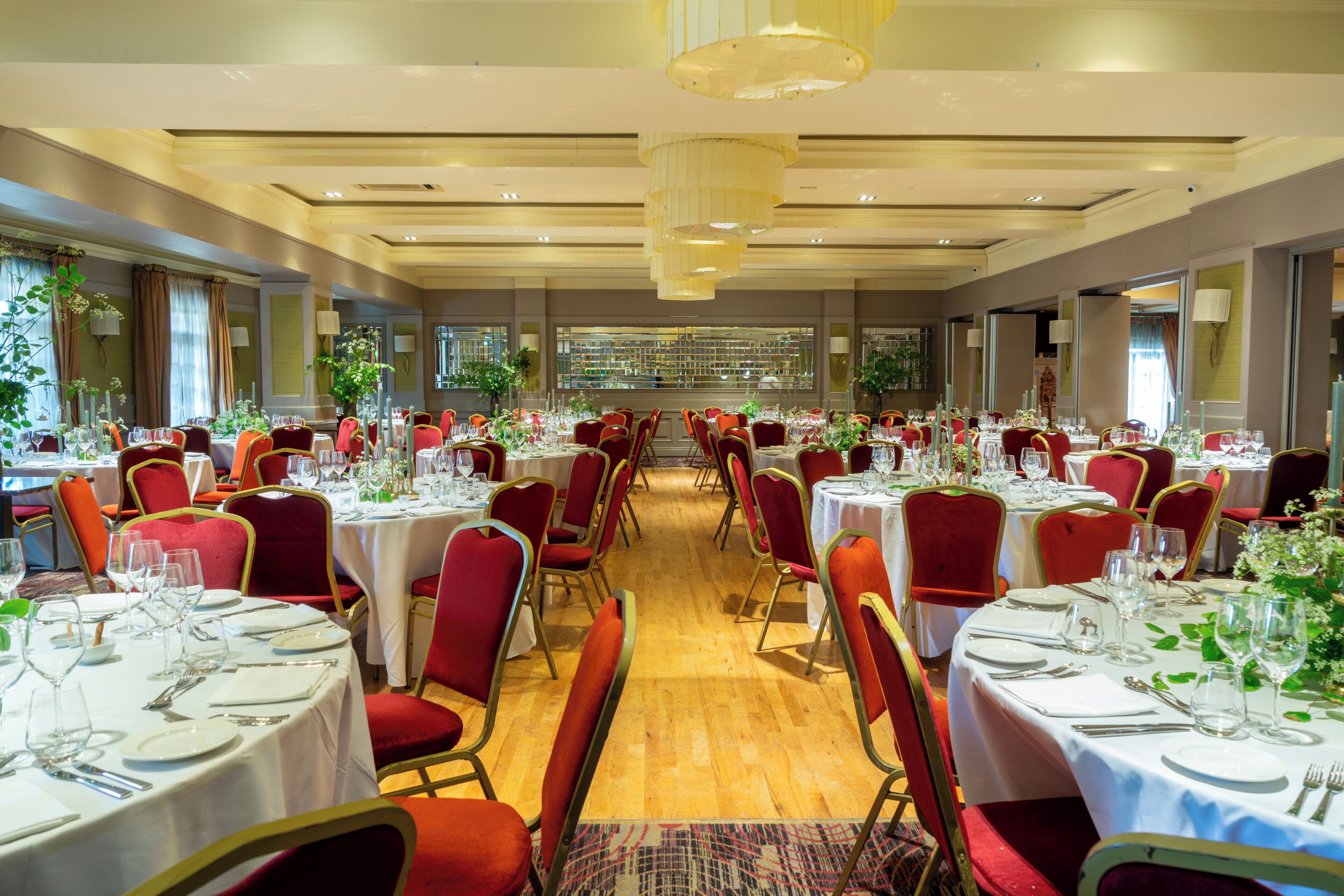 A large dining hall with multiple tables set for a formal event, featuring red chairs and white tablecloths.