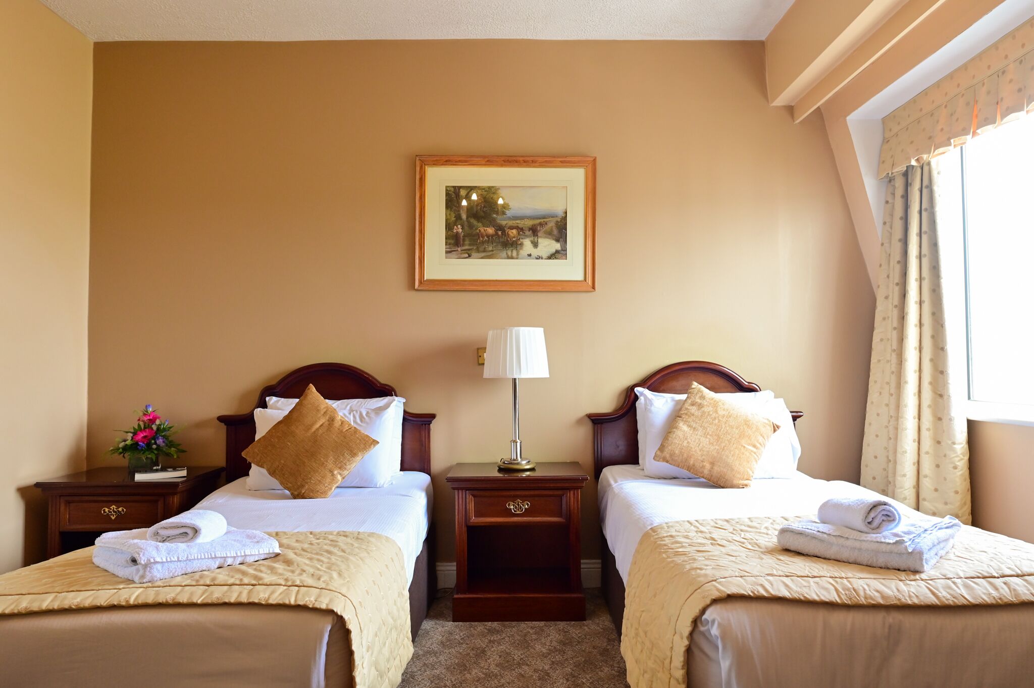 Habitación de hotel con dos camas, una mesita de noche con lámpara, un cuadro en la pared y una ventana con cortinas.