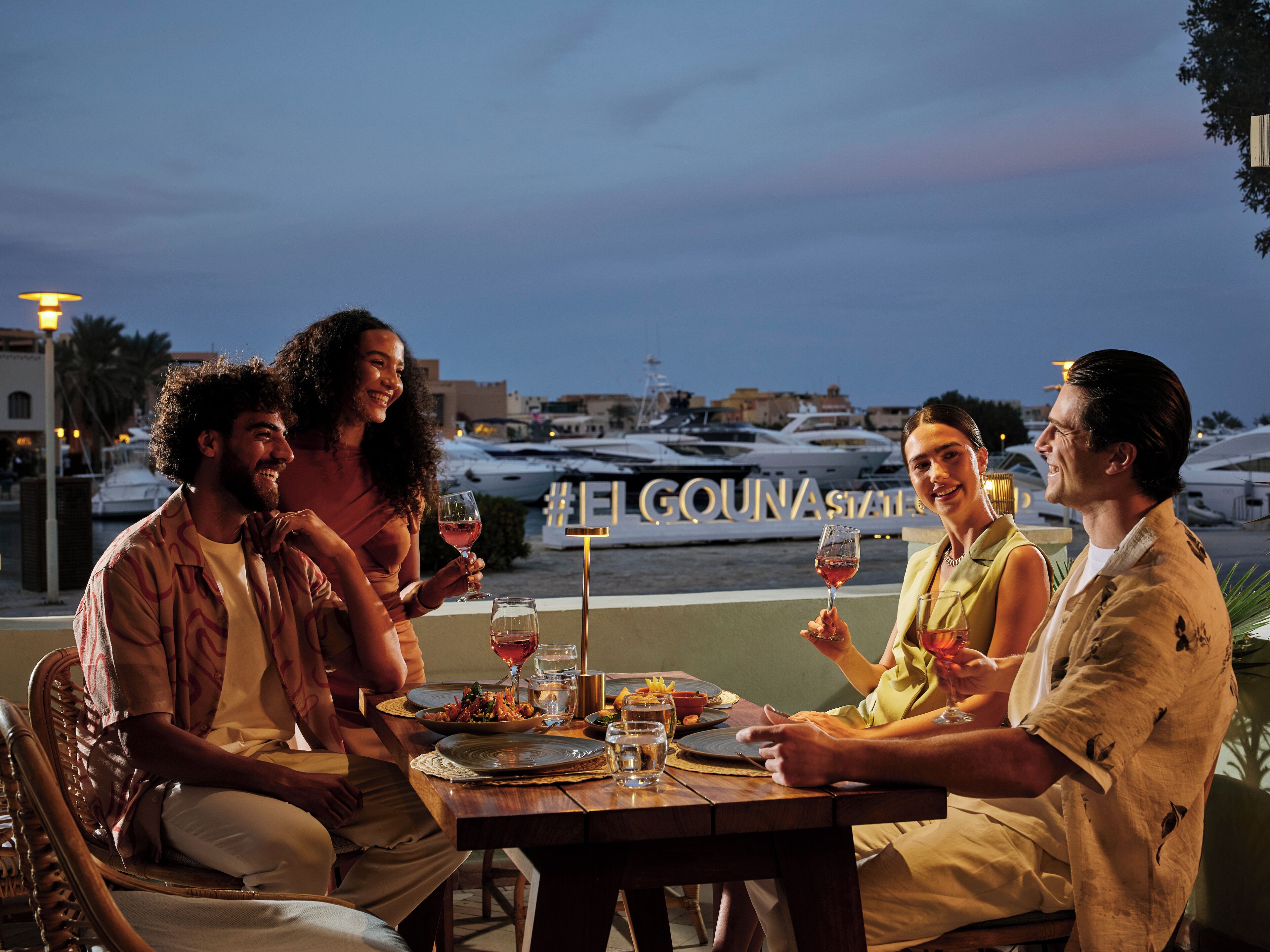 Un grupo de personas cenando en una mesa al aire libre con vista a un puerto deportivo.