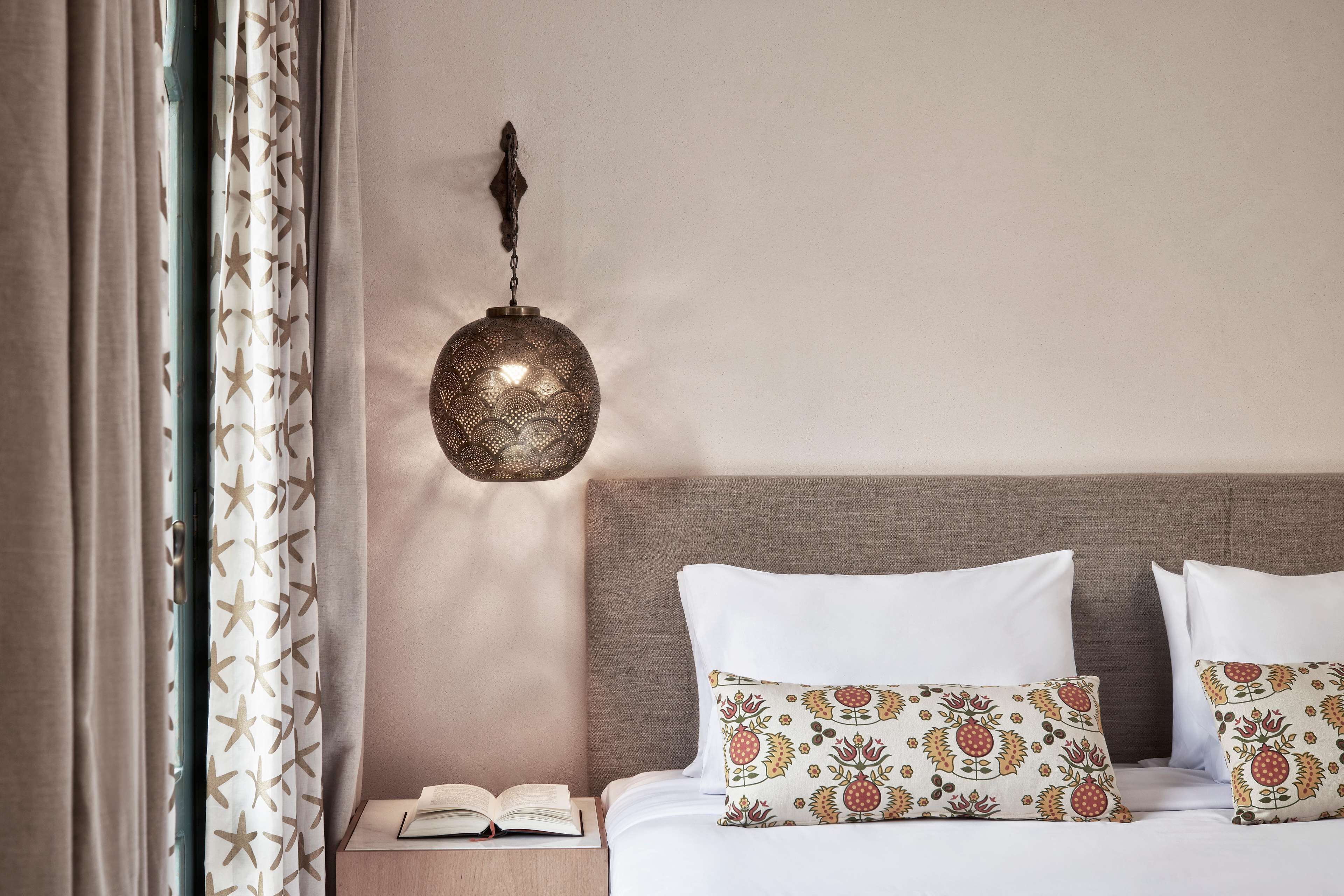 A neatly made bed with patterned pillows, a bedside table with a book, and a hanging lamp.