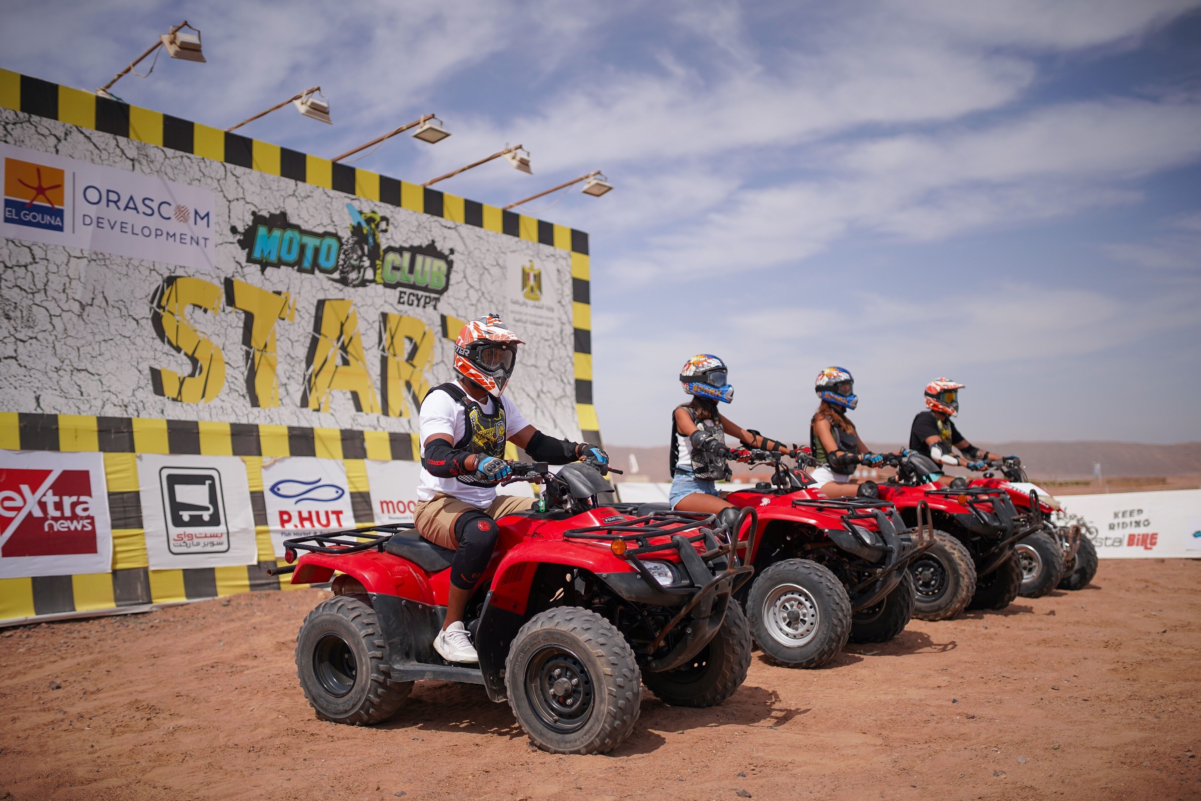 Four riders on quad bikes lined up in front of a sponsorship wall.
