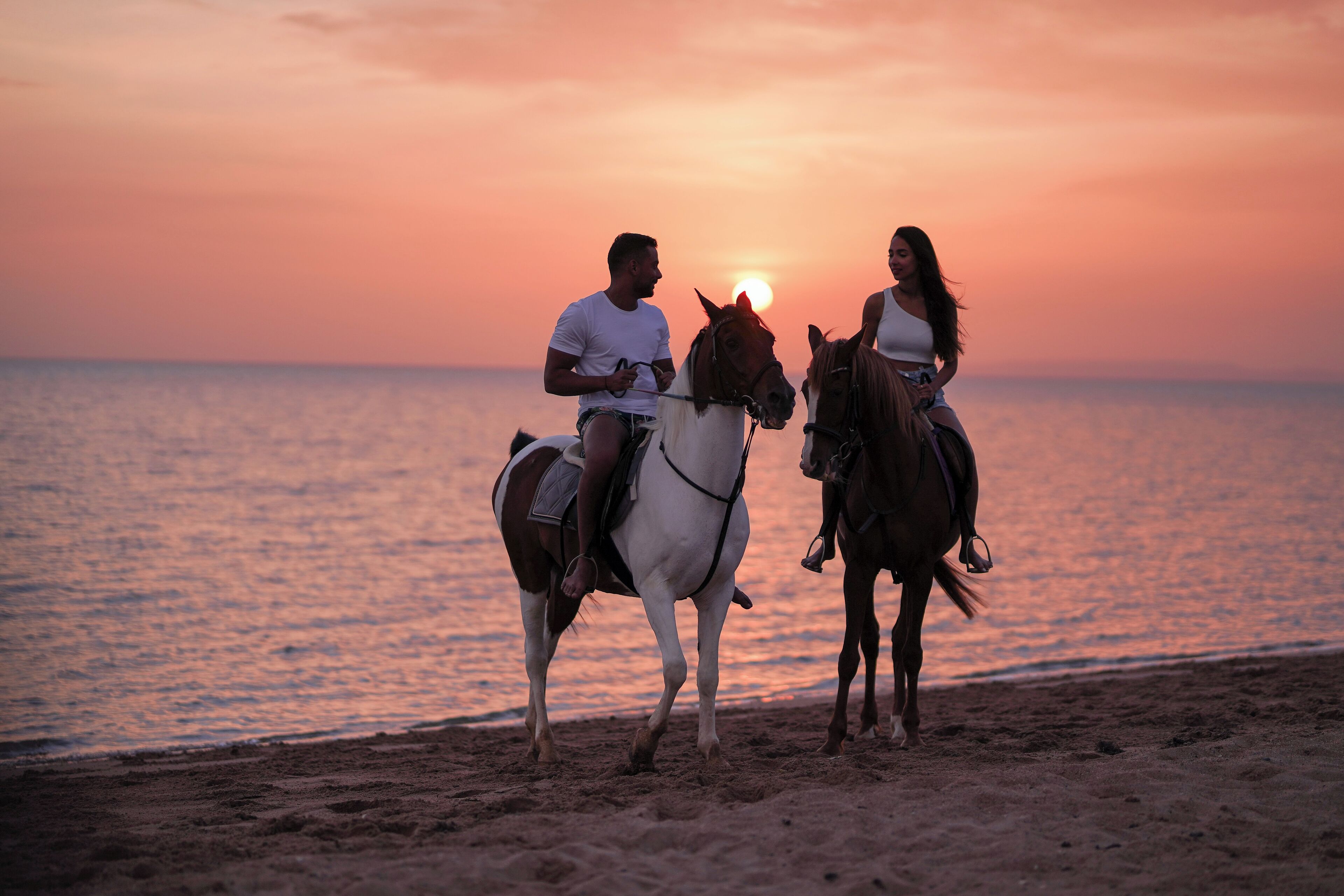 Two people riding horses on a beach at sunset.
