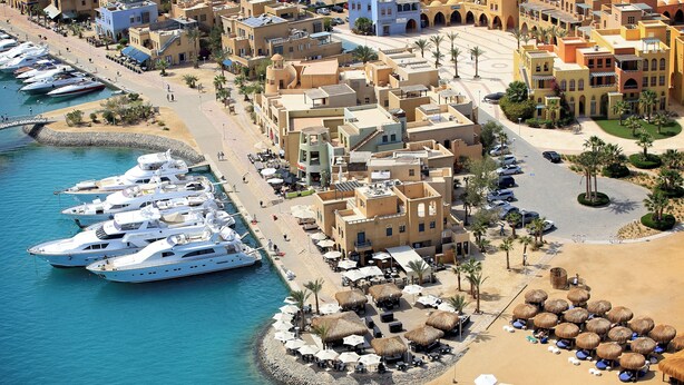 A coastal marina with yachts docked, a sandy beach with umbrellas, and a row of buildings with balconies overlooking the water. - Ali Pasha Hotel El Gouna (El Gouna)