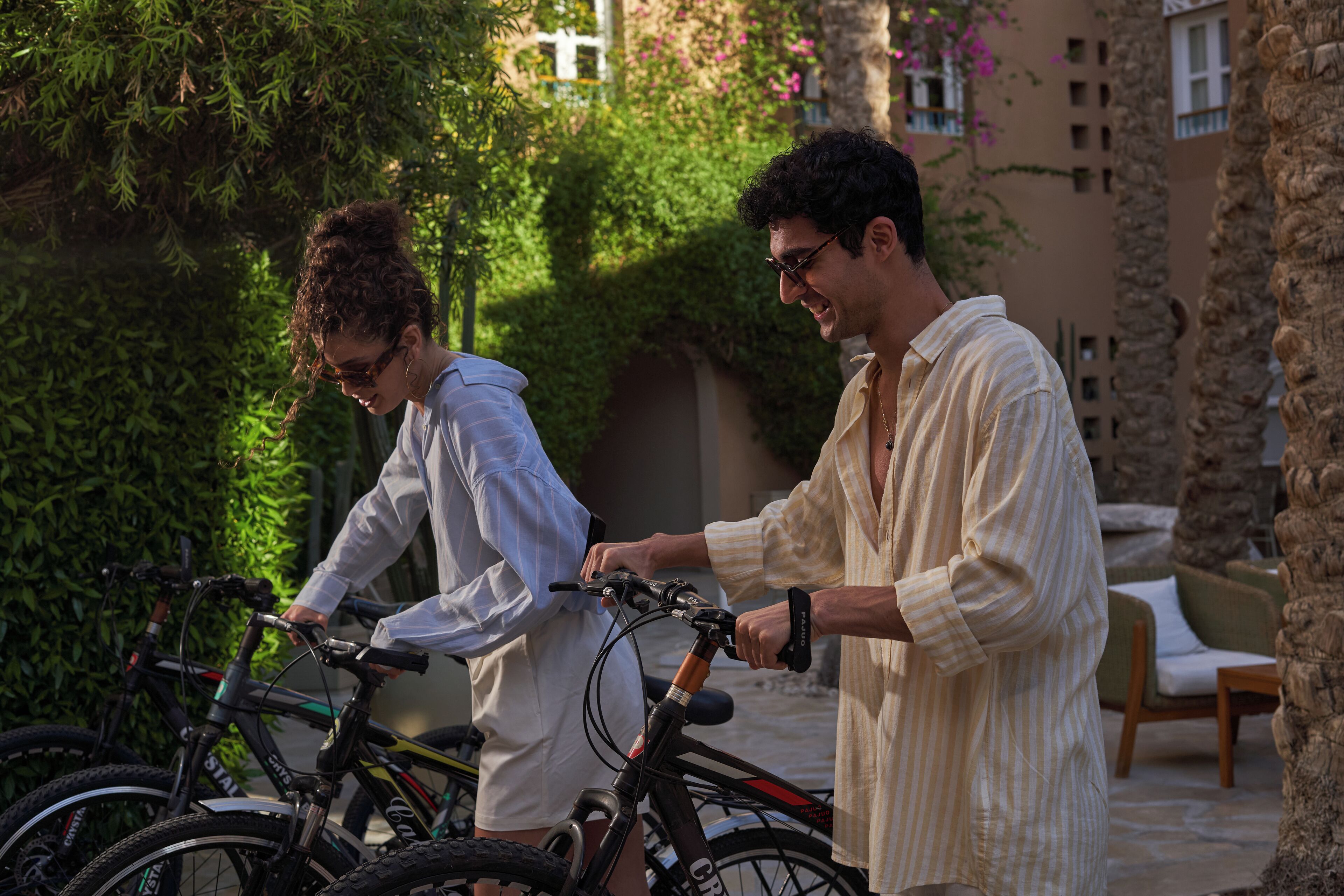 Two people standing next to bicycles in an outdoor area with greenery and stone structures.
