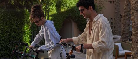 Two people standing next to bicycles in an outdoor area with greenery and stone structures.