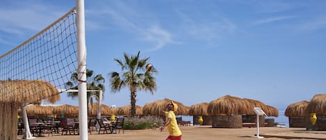 A beach volleyball court with a net, palm trees, and thatched umbrellas in the background.