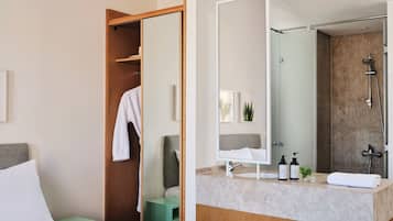 Une chambre d’hôtel moderne comprenant un lit, une table de chevet verte, une armoire en bois, une salle de bain avec douche, un miroir et un lavabo équipé de produits de toilette.