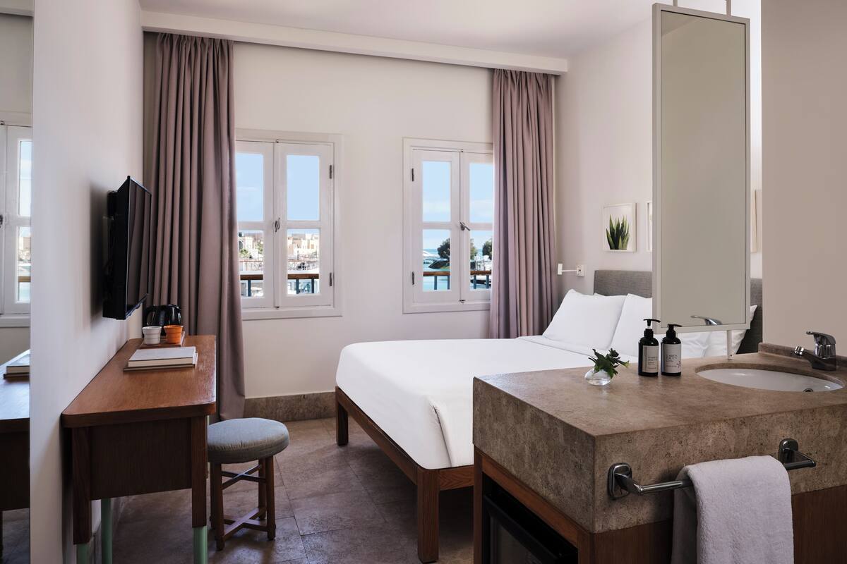 A hotel room with a bed, a desk, a TV, a bathroom with a sink and mirror, and large windows with curtains.