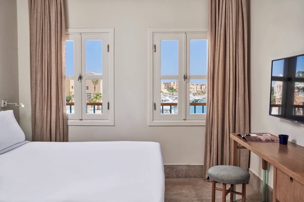 A hotel room with a bed, a desk, a chair, a TV, and windows with curtains.