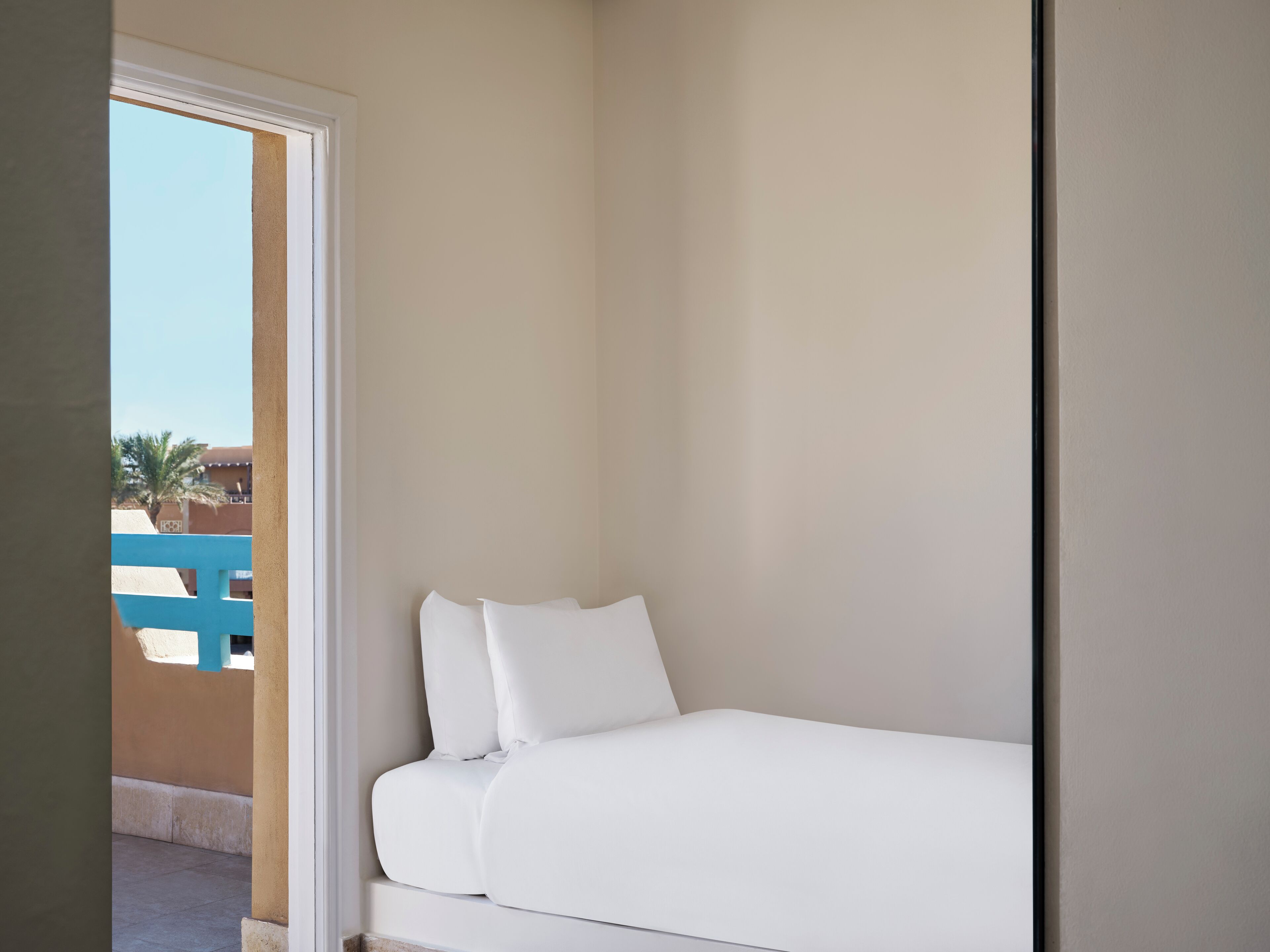 Habitación pequeña con una cama individual, paredes blancas y vistas a un edificio azul y palmeras.