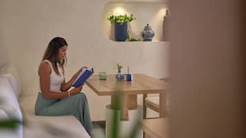 Une personne est assise à une table en bois, devant elle se trouvent un livre, un petit vase avec une plante, et un pot en céramique bleue et blanche est accroché au mur.