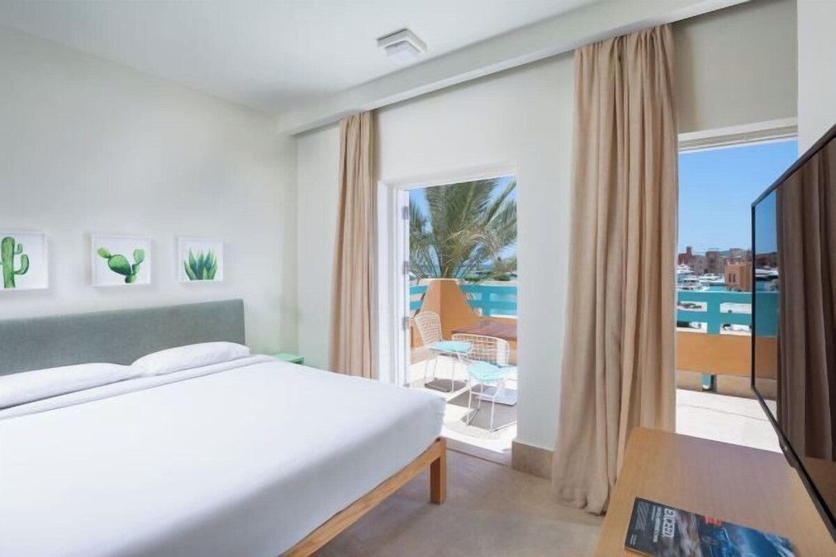 A hotel room with a bed, a television, and a balcony with a view of the city and palm trees.