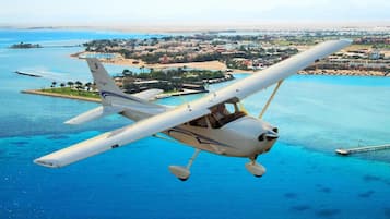 Um pequeno avião branco voando sobre uma área costeira, com vista clara para a praia e edifícios ao fundo.