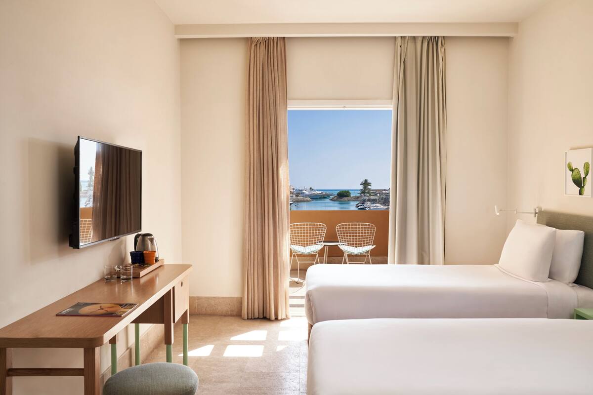 A hotel room with two beds, a desk with a TV, a coffee maker, and a view of the sea.