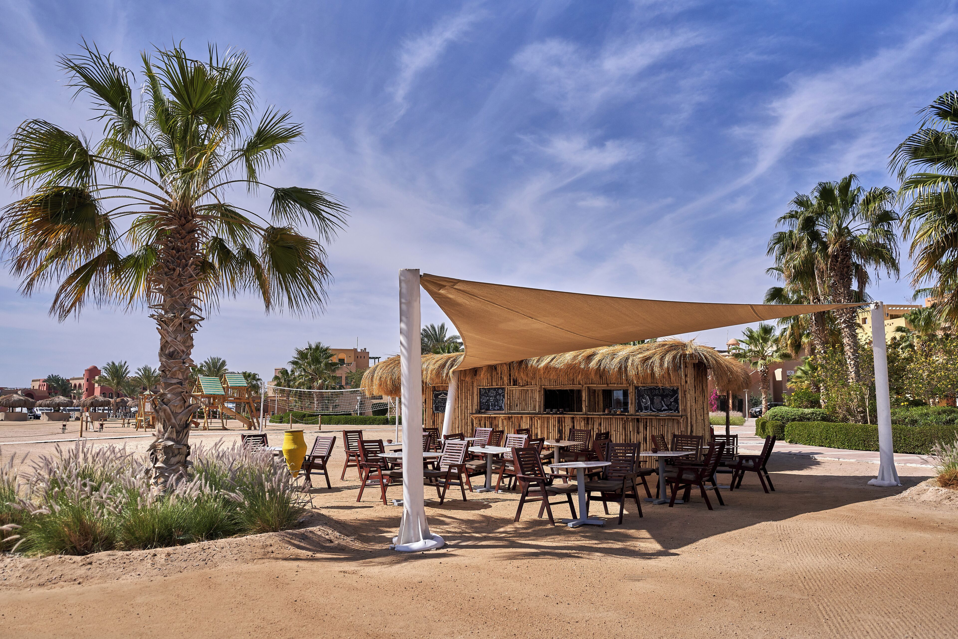 An outdoor seating area with tables and chairs, surrounded by palm trees and a thatched-roof structure.