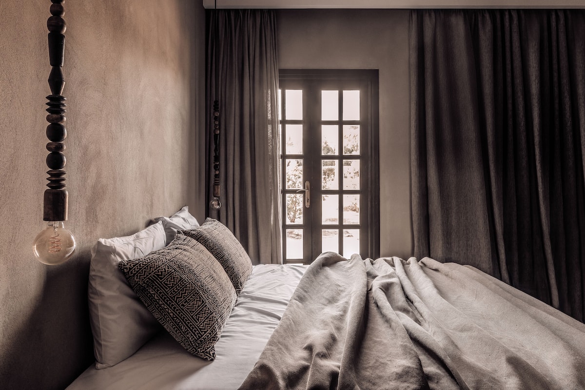 Une chambre à coucher avec un lit, des coussins à motifs, un luminaire suspendu et une fenêtre avec des rideaux.