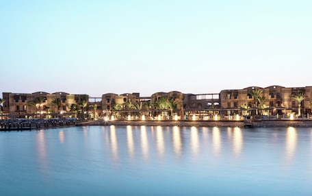 A waterfront hotel with multiple buildings, palm trees, and a calm body of water reflecting the lights.