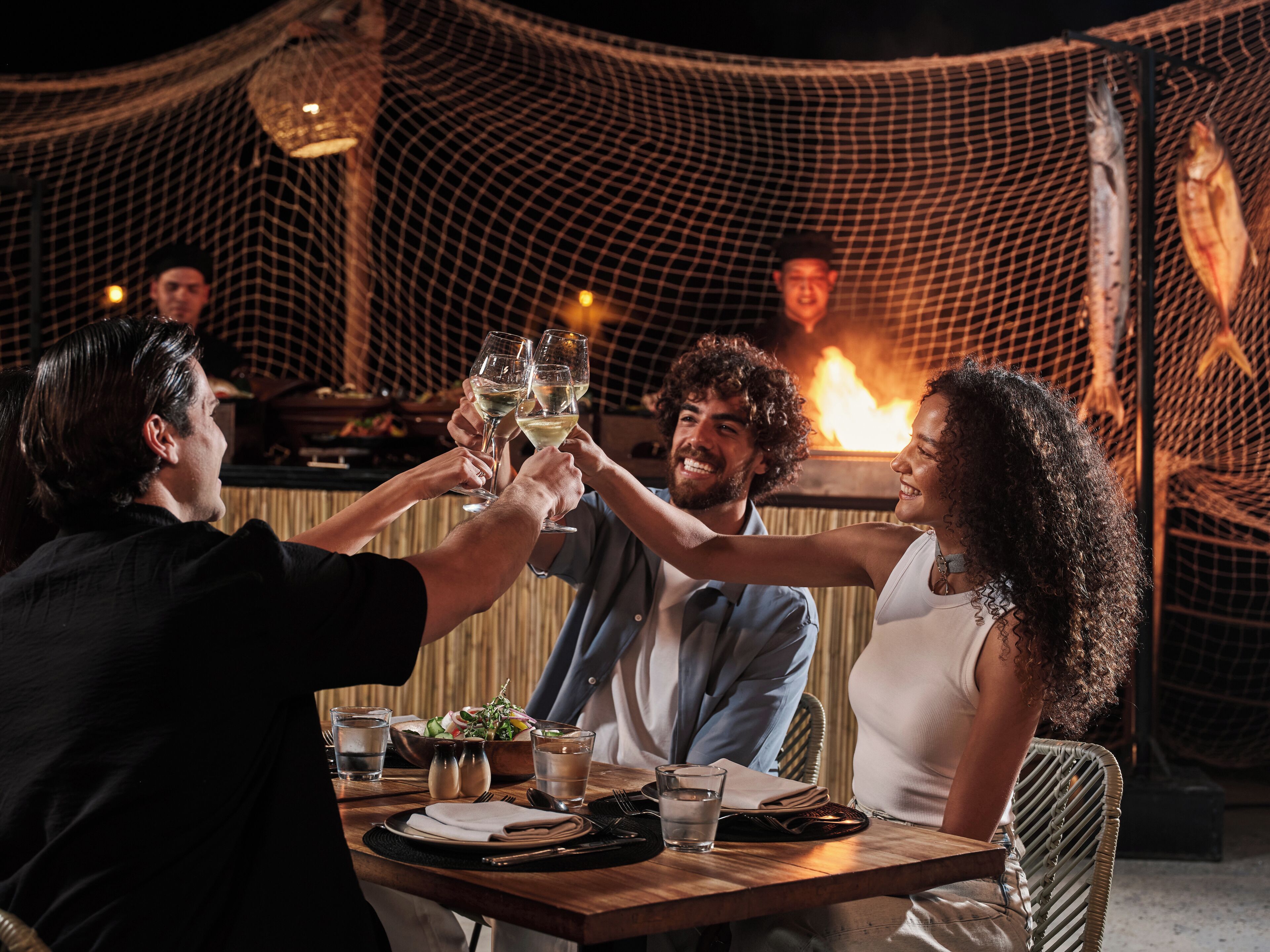 A group of people toasting with wine glasses at a wooden table with a fire in the background.