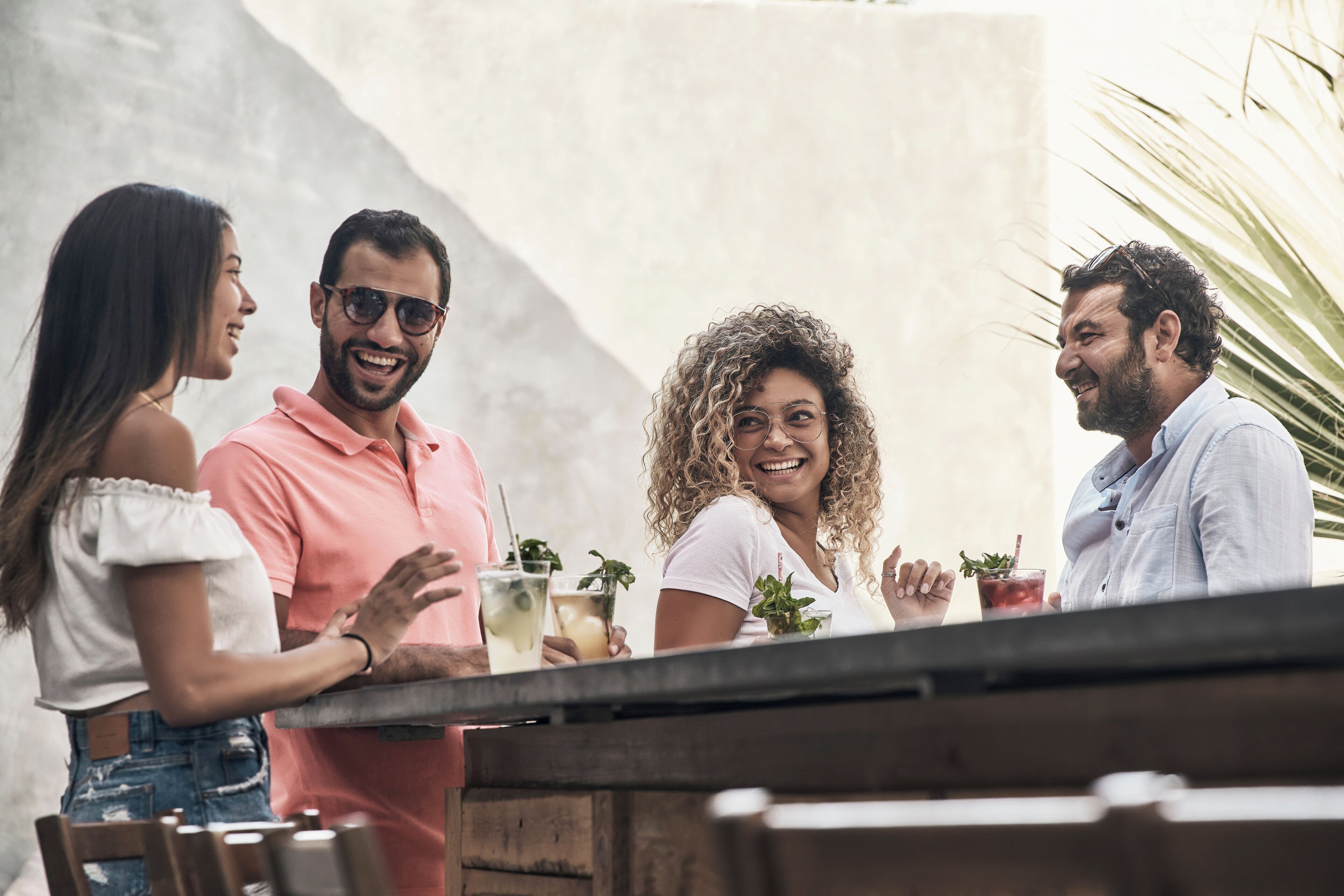 Un grupo de personas de pie junto a una mesa de madera al aire libre, sosteniendo bebidas con hojas de menta.