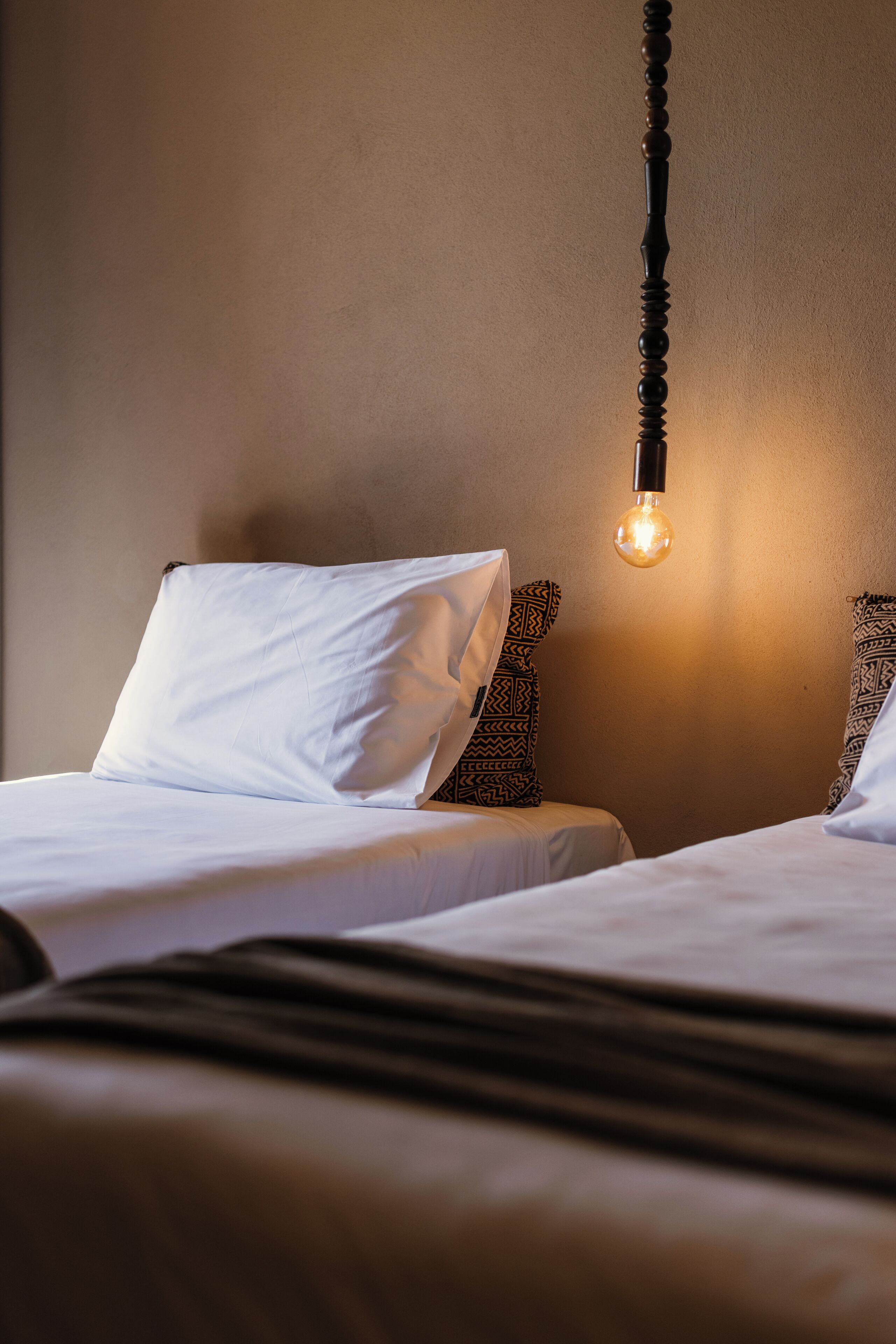 A hotel room with two beds, each with white bedding and patterned pillows. A single light bulb hangs from the ceiling, providing illumination.