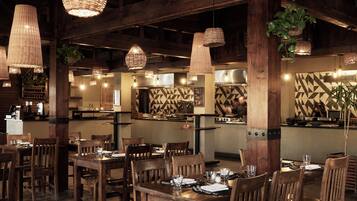 A restaurant interior with wooden tables and chairs, hanging wicker lamps, and a kitchen area with a geometric backsplash.