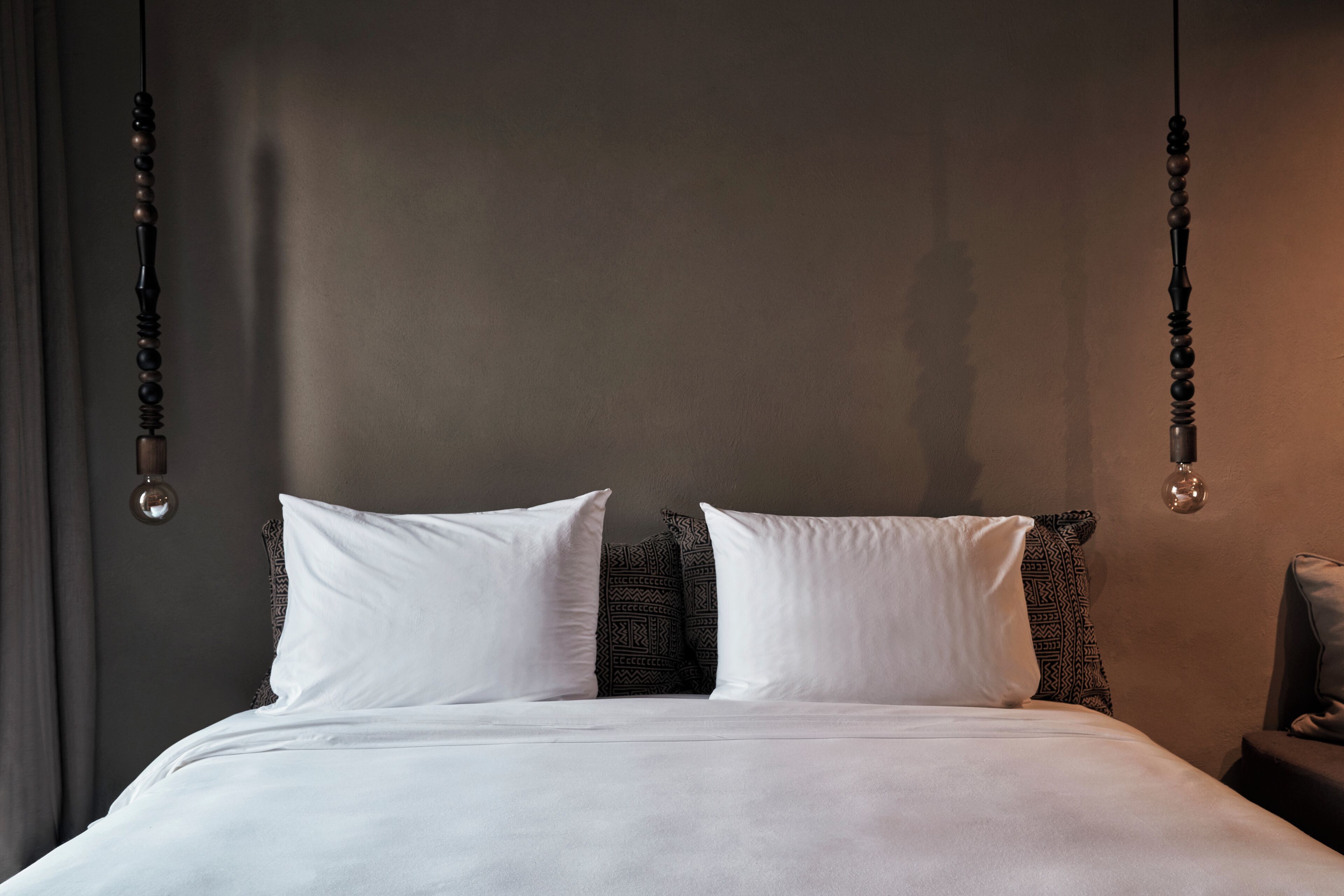 A neatly made bed with white linens and two pillows, a dark headboard, and a hanging light fixture.