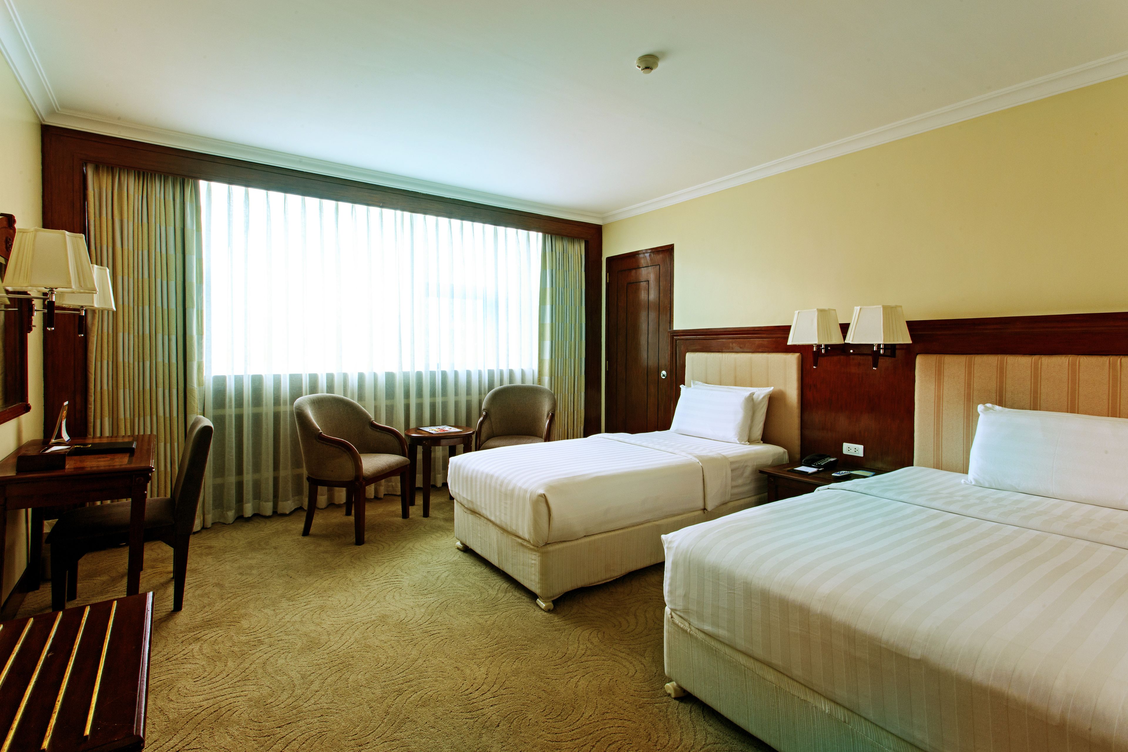 A hotel room with two beds, a desk with a chair, a small table, and a window with curtains.