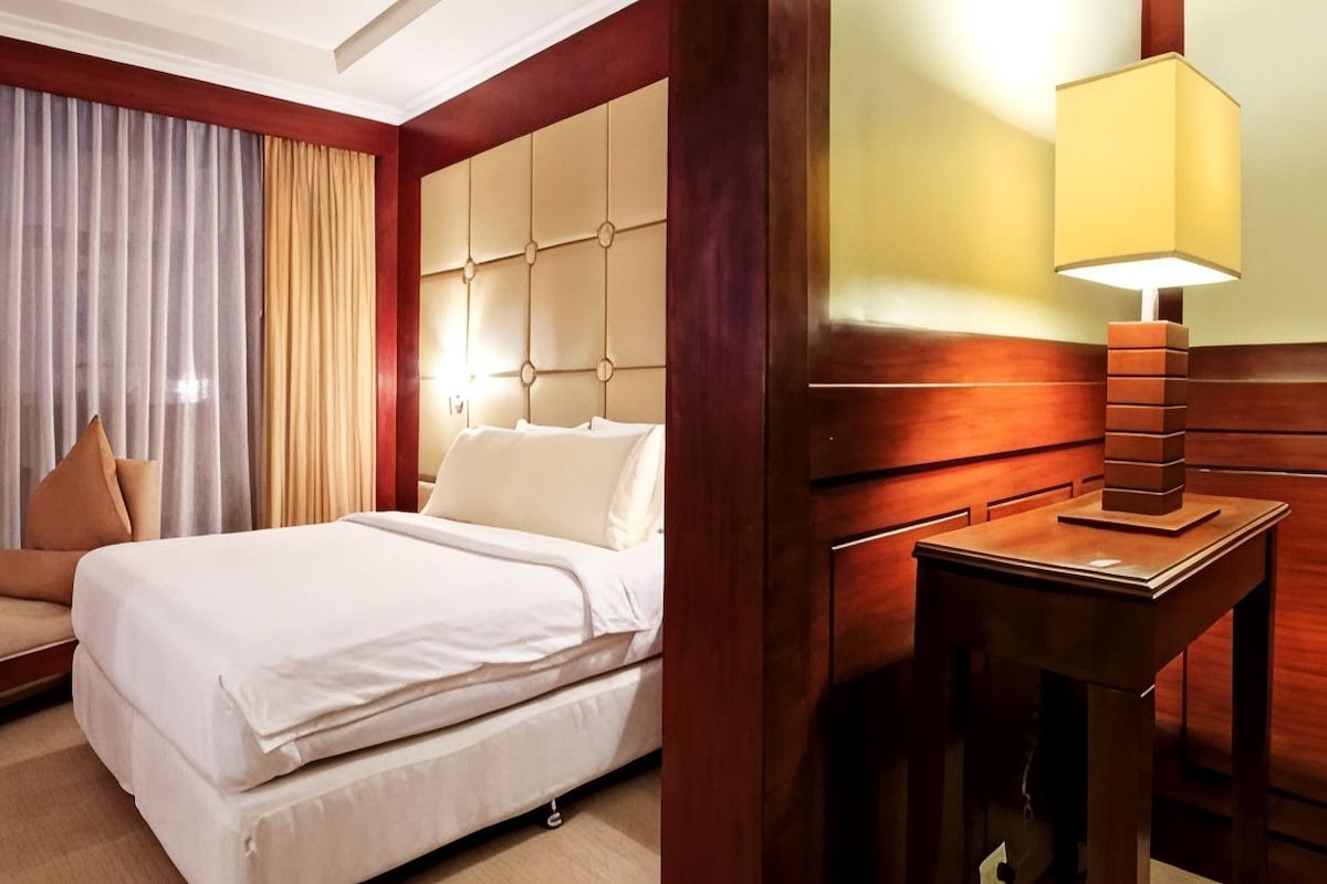 Une chambre d’hôtel avec un lit, une table de chevet avec une lampe, un canapé et une grande fenêtre avec des rideaux.