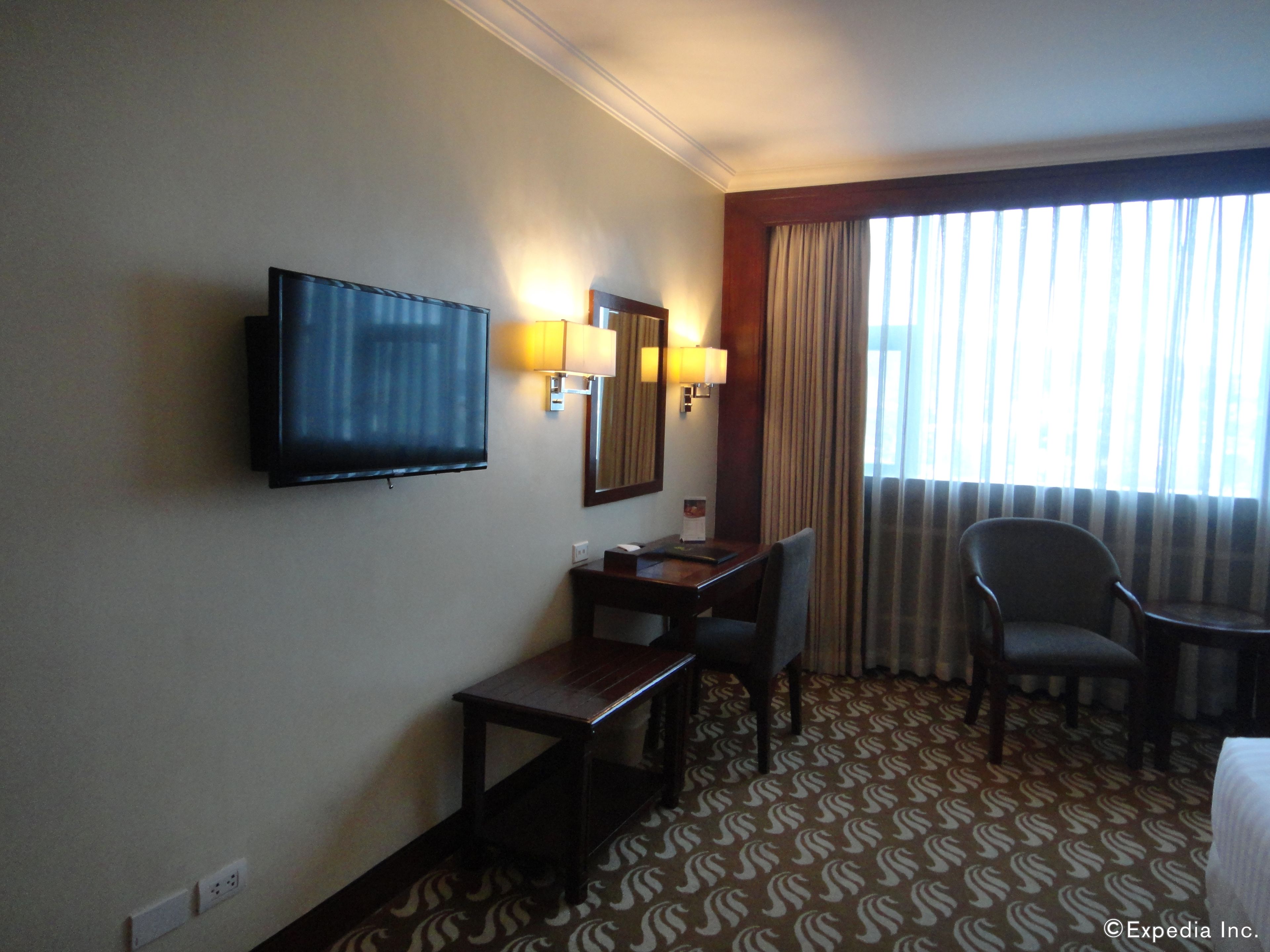 Habitación de hotel con televisión de pantalla plana, escritorio con silla, cama y ventana grande con cortinas.