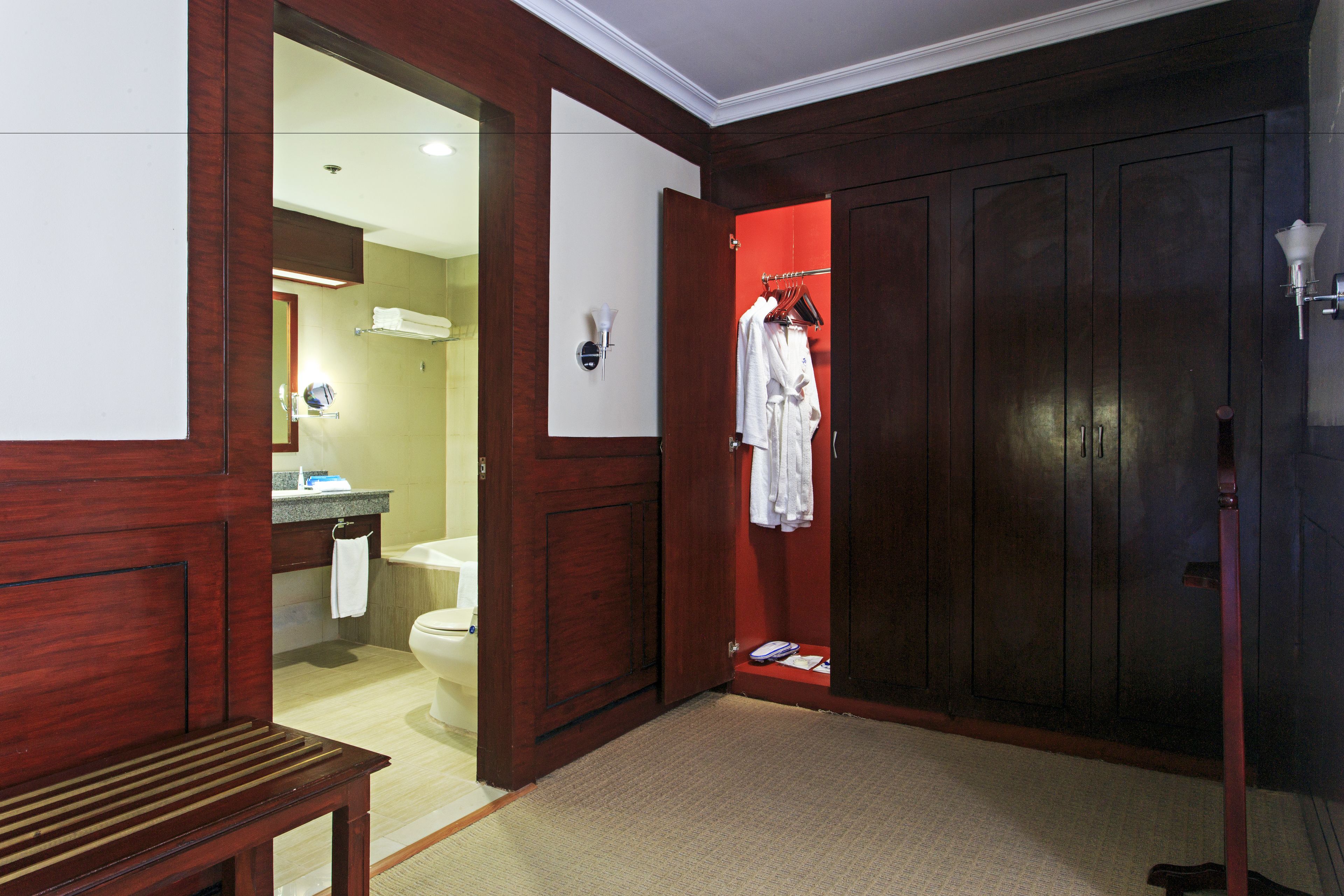 A hotel room with a wooden wardrobe, a bench, a red door, and a bathroom visible through an open door.