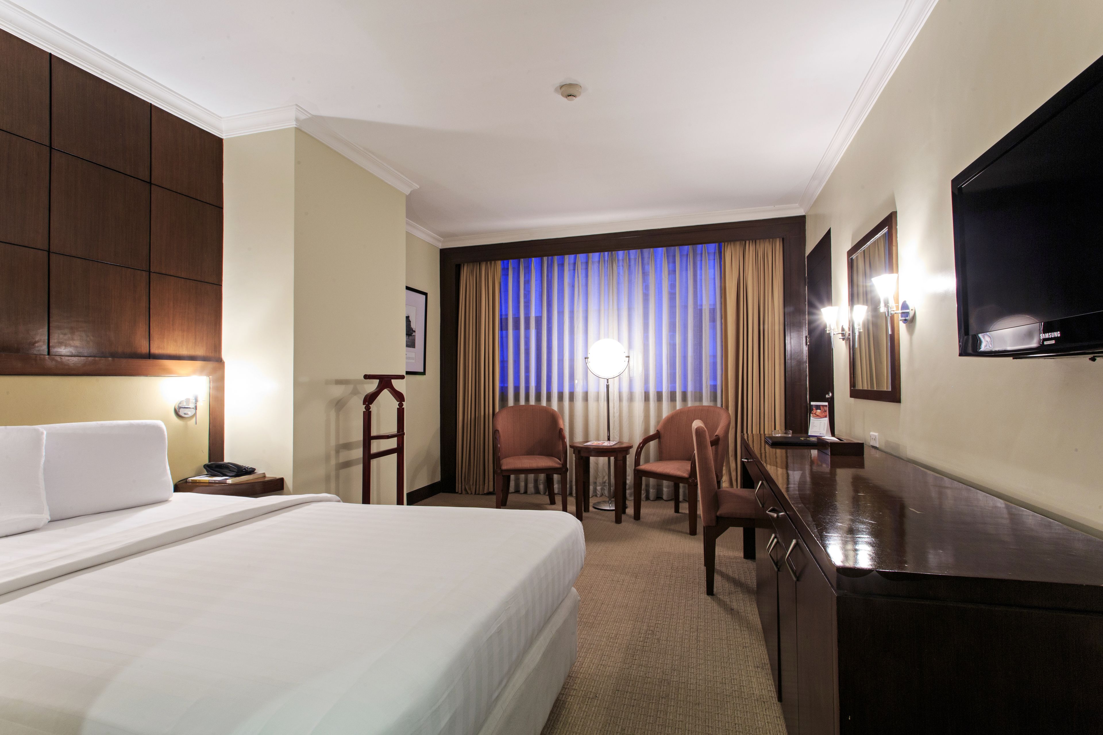 A hotel room with a large bed, a desk, two chairs, a TV, and a window with curtains.