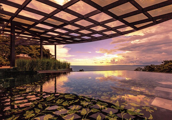 An infinity pool with a view of the sunset over the ocean, surrounded by a wooden pergola and lush greenery.