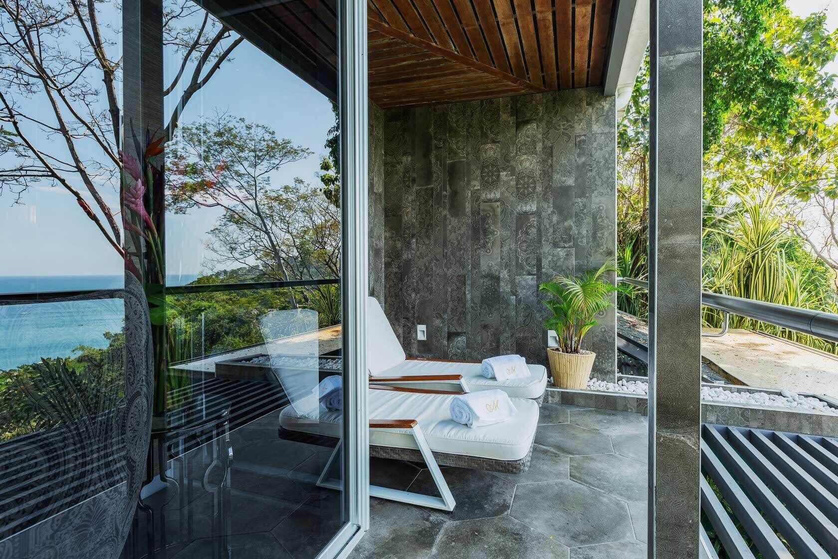 A modern outdoor seating area with a view of the sea, featuring a glass partition, a concrete wall, and a wooden ceiling.