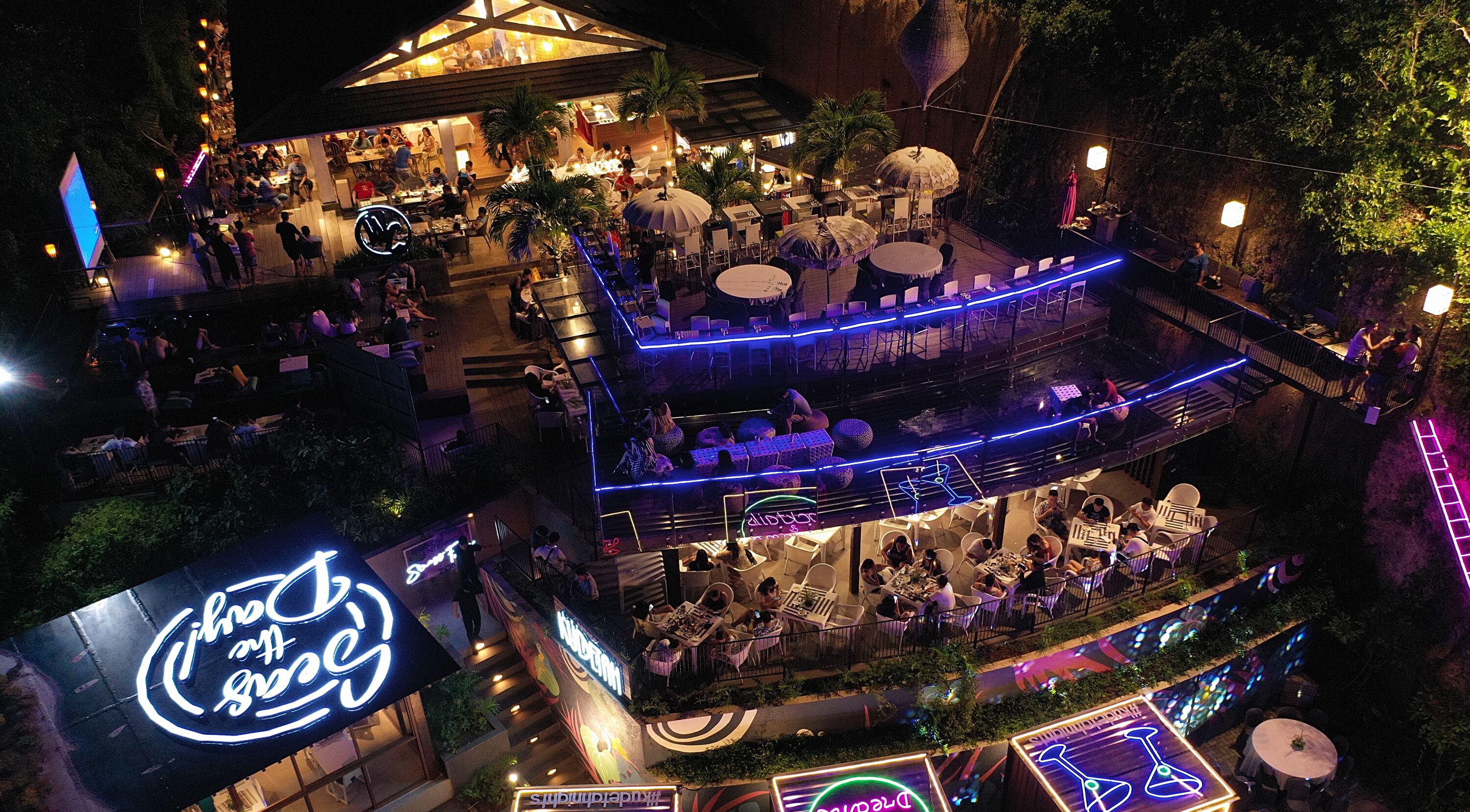 An outdoor dining area with multiple levels, illuminated by blue and purple lights, surrounded by lush greenery.