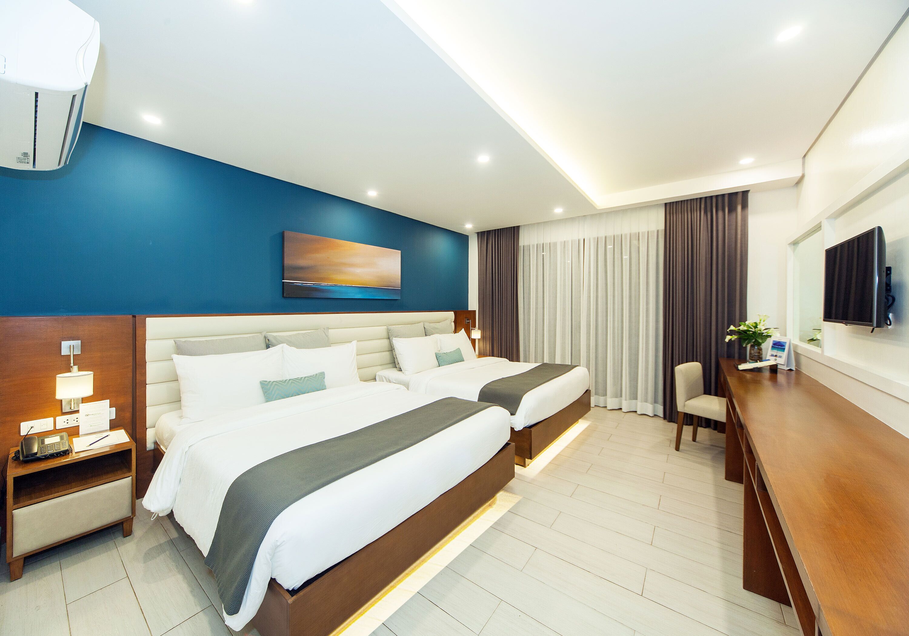 Ein modernes Hotelzimmer mit zwei Betten, einem Schreibtisch, einem Sessel, einem Fernseher und einer Klimaanlage.