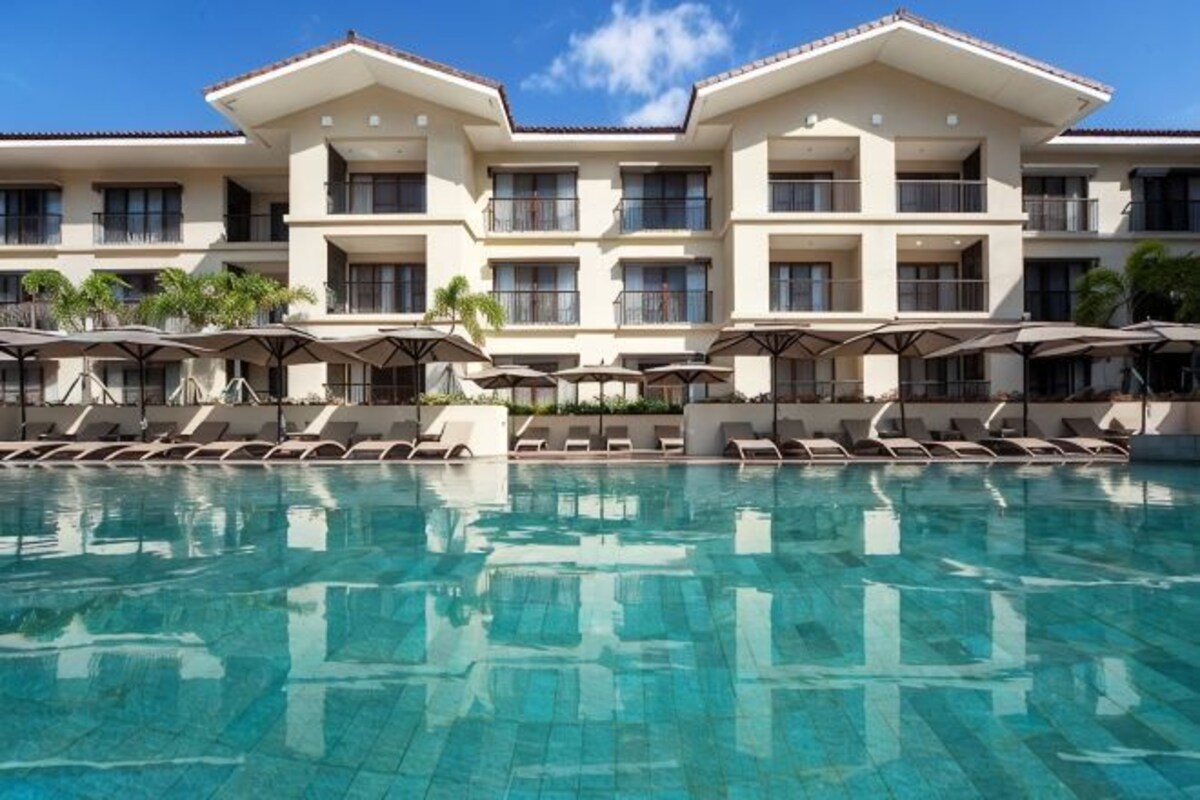 Un hotel con piscina, tumbonas, sombrillas y un edificio de varios pisos con balcones.