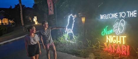 Two people standing near a neon sign that reads "Welcome to the Neon Night Safari."