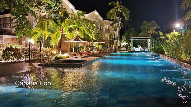 A nighttime view of a Cabana Pool with a clear pool area, lounge chairs, and a building with a lighted facade in the background.