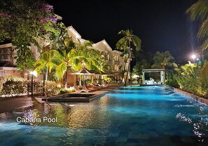 Vue nocturne d’une piscine de type « cabana », avec une zone de baignade dégagée, des chaises longues et un bâtiment dont la façade est illuminée en arrière-plan.
