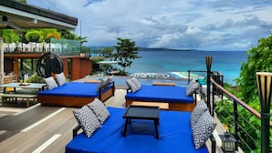 A rooftop lounge with blue cushioned seating, a pool, and a view of the ocean.