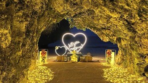 An illuminated heart-shaped sign with the words "Will you marry me?" inside an archway, surrounded by flowers and candles.