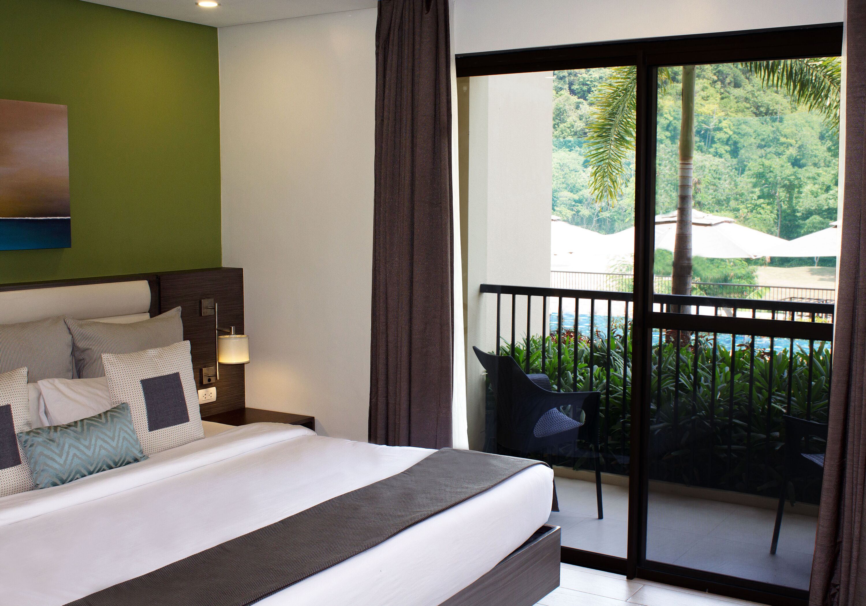 A hotel room with a large bed, a balcony with a view of a pool and greenery.