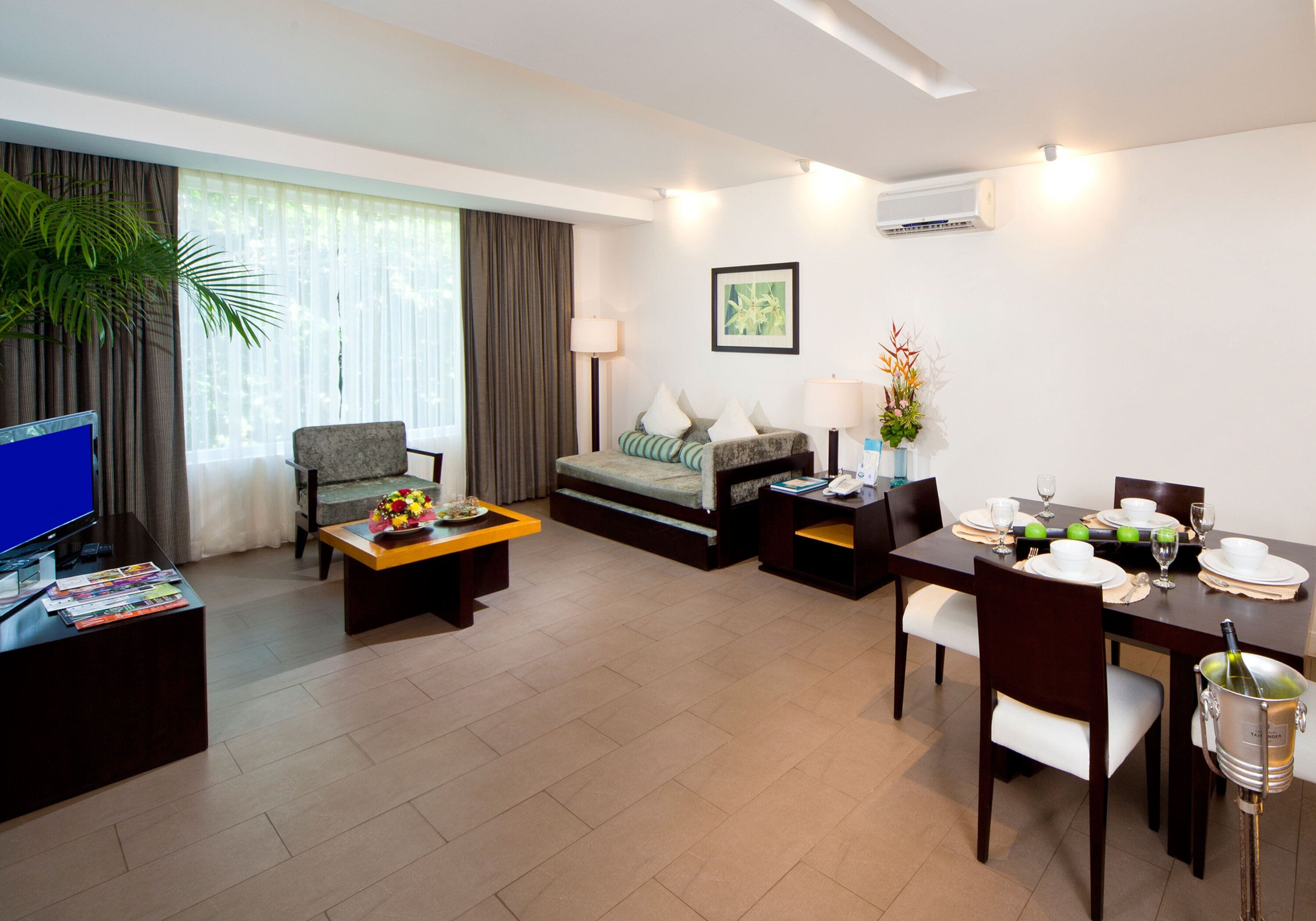 A modern hotel room with a flat-screen TV, a sofa, a dining table set for two, and a minibar.