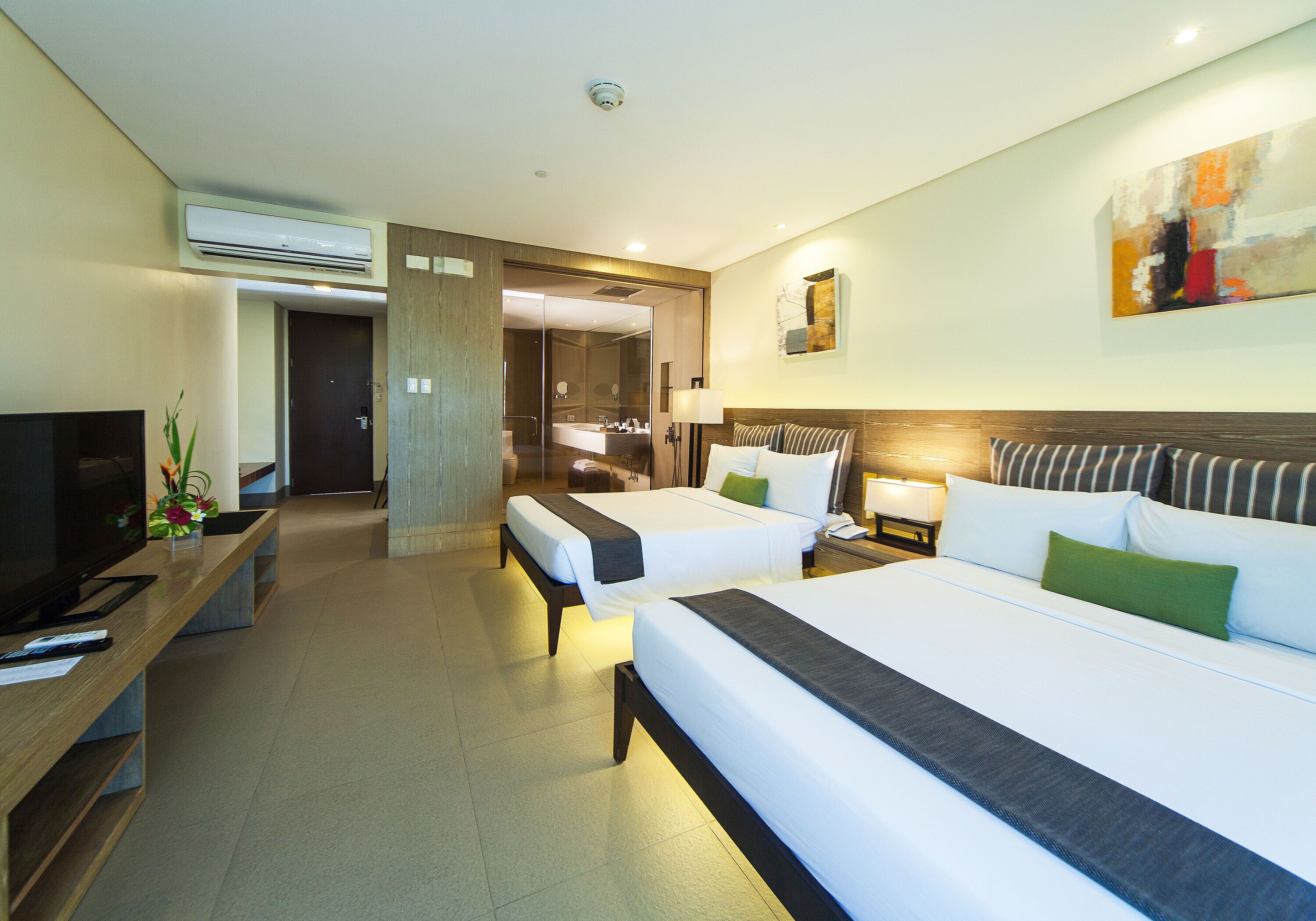 A hotel room with two beds, a TV, a desk, and a bathroom visible through a glass partition.