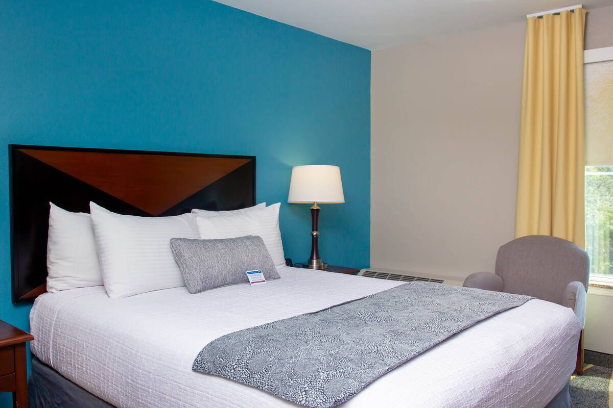 A neatly made bed with a headboard, a bedside lamp, and a chair by a window with curtains.