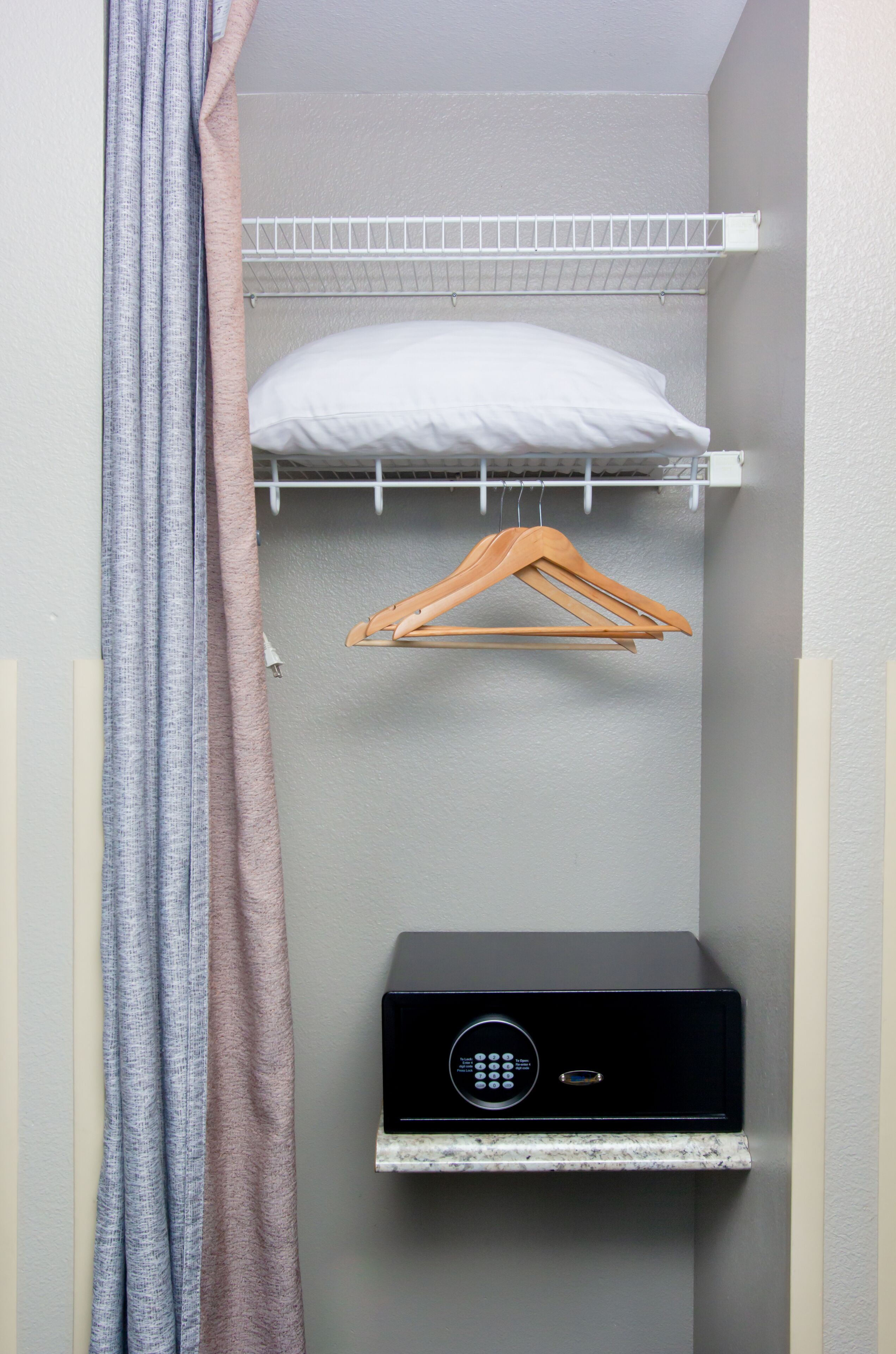 A small, enclosed room with a white shelf, a hanging wooden hanger, a white pillow, and a black electronic device on a marble shelf.