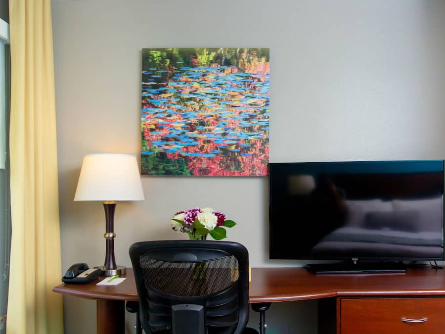 A hotel room with a desk, a lamp, a telephone, a TV, and a painting on the wall.