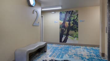 Habitación de hotel con un cartel con el número 2, un banco, un cuadro abstracto y colorido en la pared, y una alfombra azul estampada.