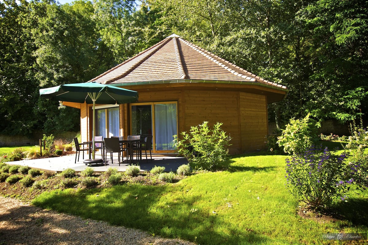 A wooden cabin with a tiled roof, a patio with outdoor furniture, and a garden with shrubs and flowers.